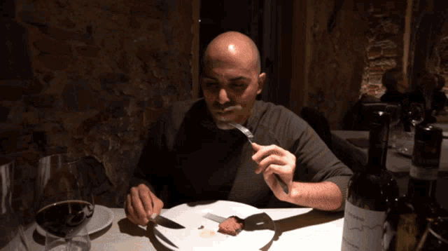 a man is sitting at a table eating a piece of meat with a bottle of wine in front of him