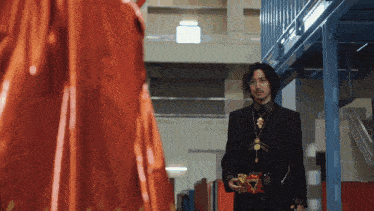 a man in a black suit and gold necklace is standing in a building