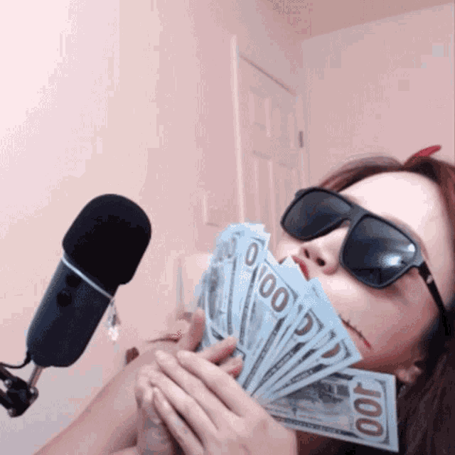 a woman wearing sunglasses is holding a fan of 100 dollar bills in front of a microphone