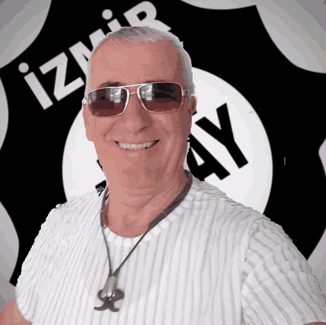 a man wearing sunglasses and a necklace smiles in front of an izmir ay logo