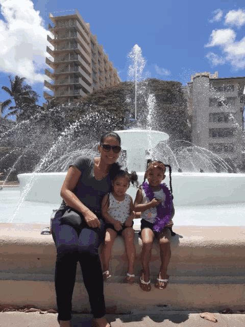 a woman sits next to two little girls in front of a water fountain