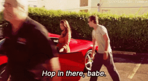 a man and a woman are standing next to a red sports car and the man says hop in there babe