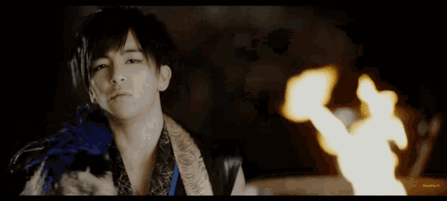 a young man with a blue feather on his shoulder stands in front of a fire