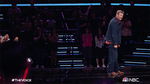 a man in a denim jacket and plaid shirt stands on a stage in front of a nbc audience