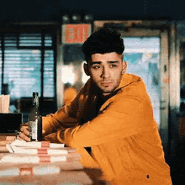 a man in a yellow hoodie is sitting at a table with a bottle of alcohol in his hand .