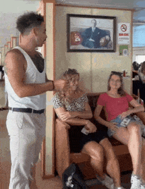 a man in a white tank top talks to two women sitting on a couch