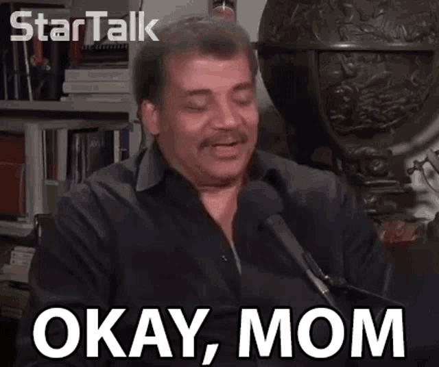 a man in a black shirt is talking into a microphone with the words okay mom above him