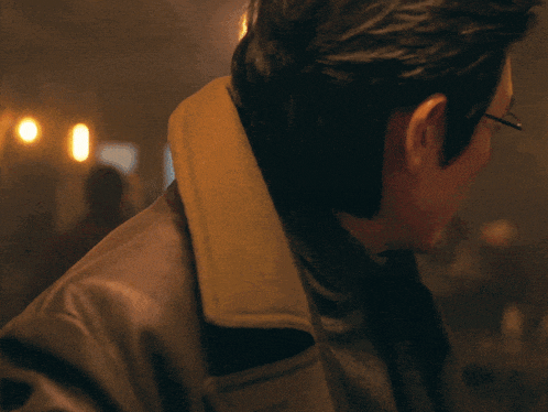 a man wearing glasses and a trench coat looks at the camera