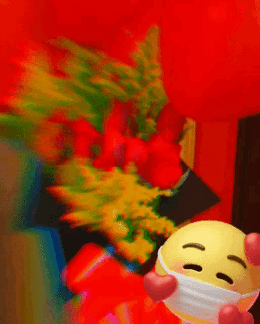 a yellow smiley face wearing a mask and holding a heart in front of a bouquet of flowers