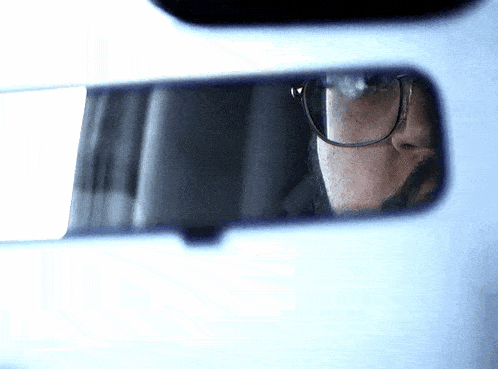 a man wearing glasses looks at himself in the rear view mirror