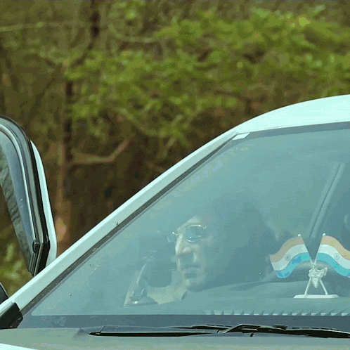 a man driving a white car with a small indian flag on the windshield