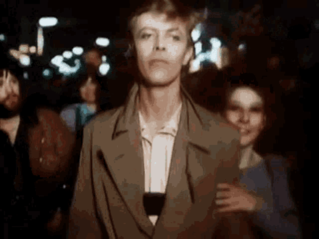 a man in a trench coat is standing in a crowd of people at night .