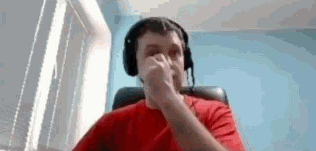 a man wearing headphones and a red shirt is sitting in a chair covering his nose .