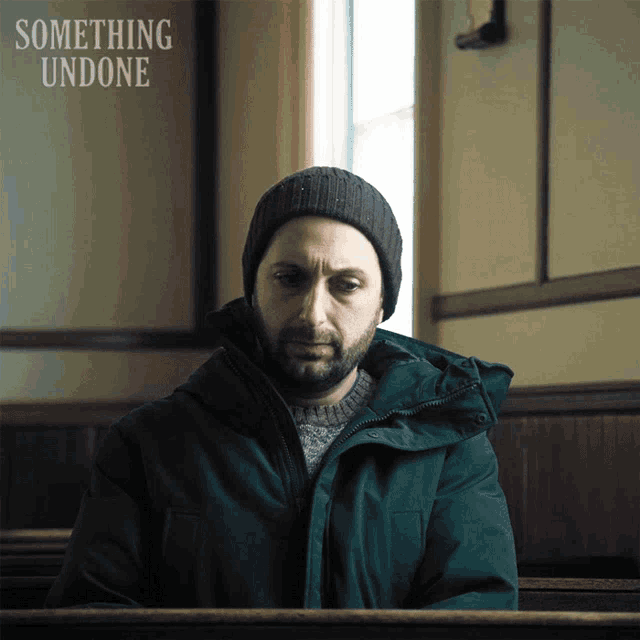 a man in a green jacket sits in a church with something undone written on the wall behind him