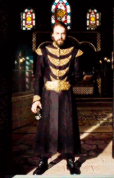 a man in a black and gold coat is standing in front of a stained glass window .