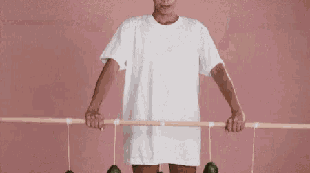 a woman in a white t-shirt is holding a wooden stick with avocados hanging from it