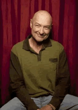 a bald man is wearing a green and black shirt and standing in front of a red curtain .