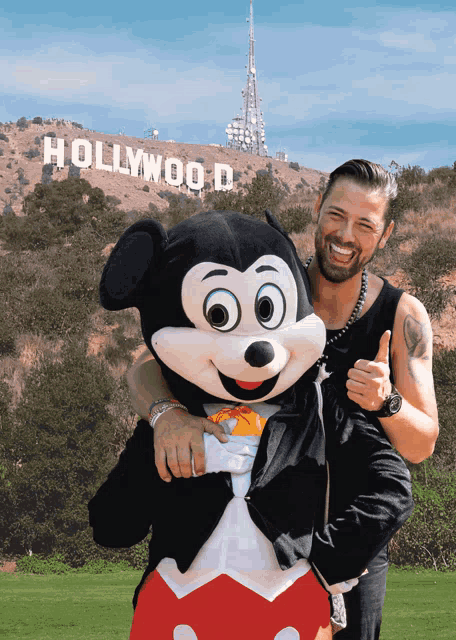 a man is holding a mickey mouse mascot in front of a hollywood sign