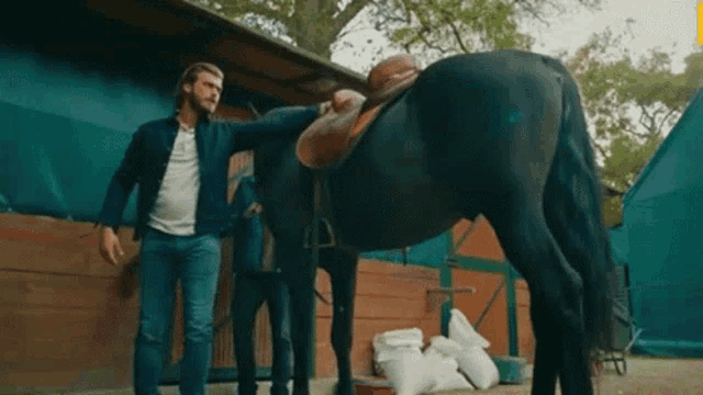 a man is standing next to a horse with a saddle on it in a stable .