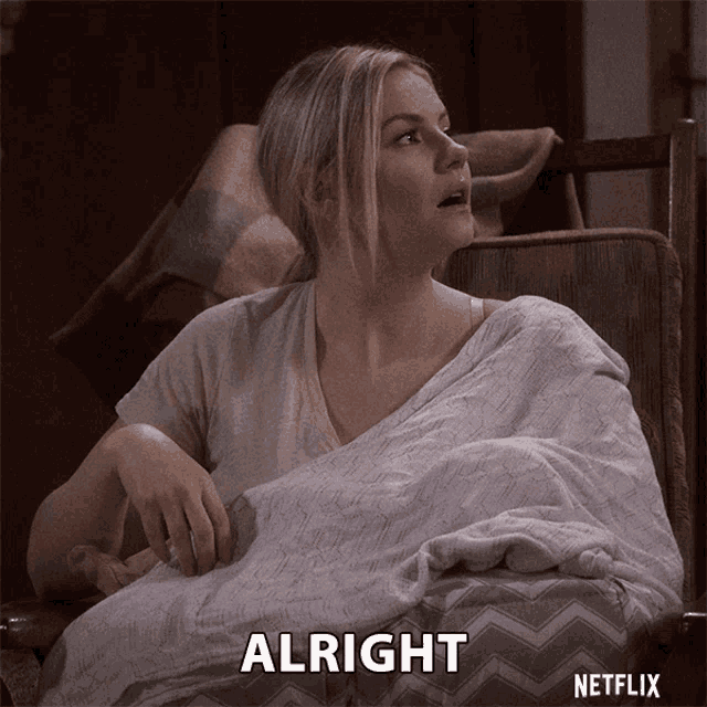 a woman is sitting in a chair with a blanket on her shoulders and the word alright is on the screen behind her