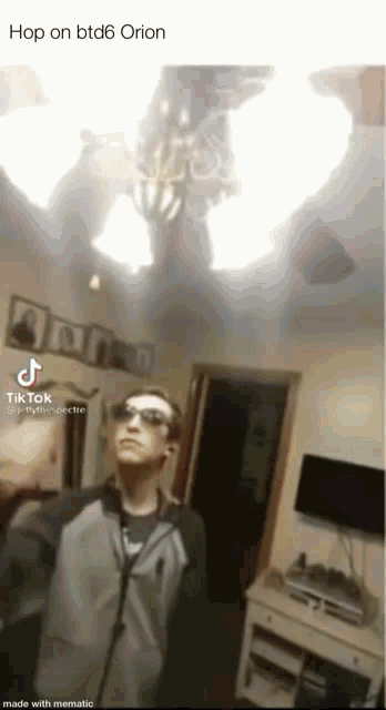 a man wearing sunglasses is standing in a living room with a chandelier hanging from the ceiling .