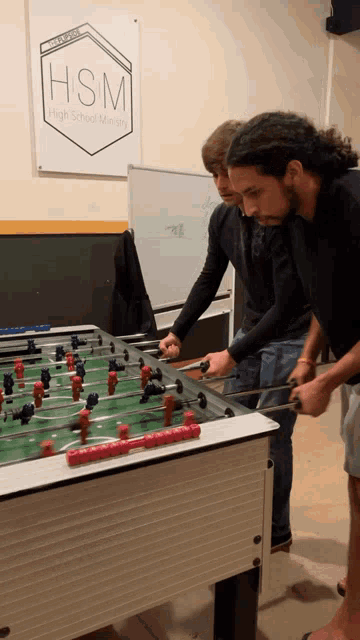 three men are playing a game of foosball in front of a sign that says hsm