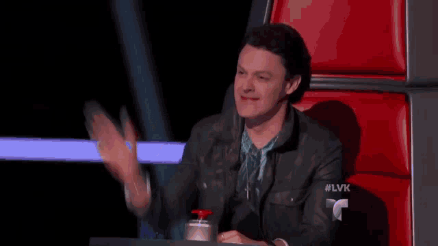 a man in a leather jacket is sitting in a chair and waving his hand .