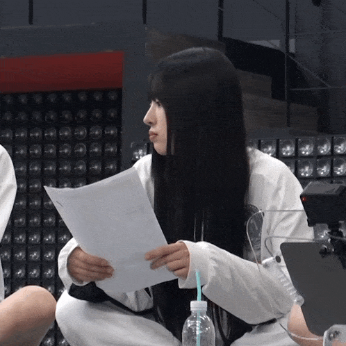 a woman with long black hair is reading a piece of paper next to a bottle of water