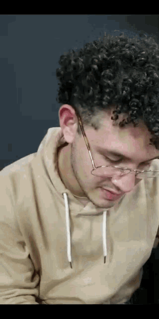 a young man with curly hair wearing glasses and a tan sweatshirt