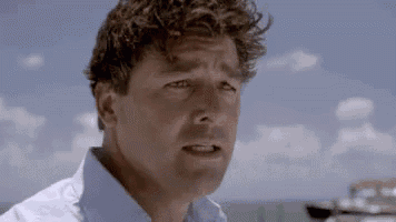a man with curly hair is standing on a beach with boats in the background and making a funny face .