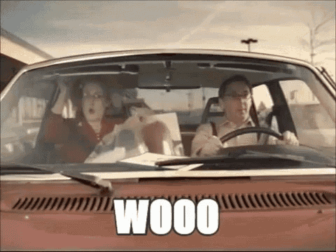 a man and a woman are driving a red car and the word wood is on the hood .
