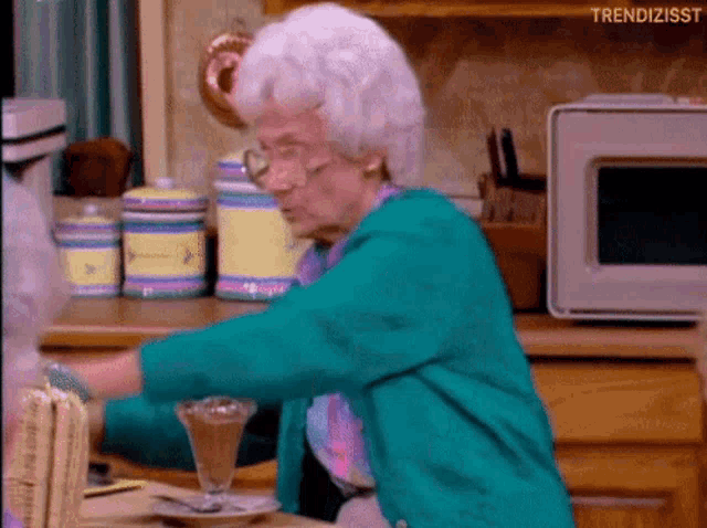 an elderly woman in a green jacket is sitting at a table with a glass of chocolate pudding .