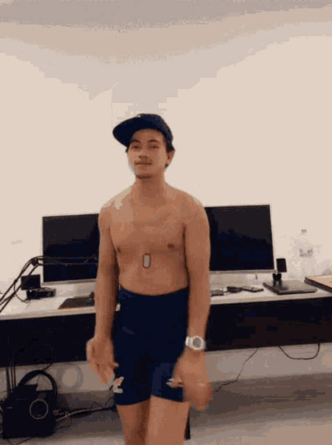 a shirtless man wearing a hat and shorts stands in front of a computer monitor