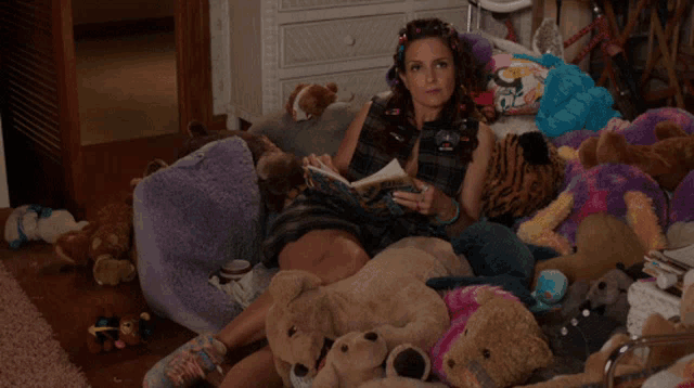 a woman is laying on a couch surrounded by stuffed animals and reading a book