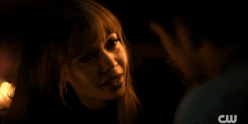 a woman looks at a man in a dark room with a cw logo behind her