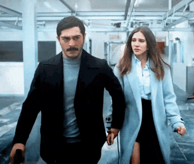 a man in a black jacket and a woman in a blue coat are walking down a hallway holding hands
