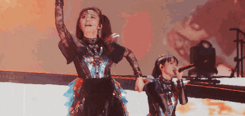 two women are singing into microphones on a stage with their arms in the air .