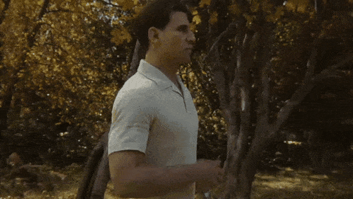 a man in a white shirt is standing in a park with his hands on his chest