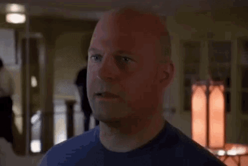 a bald man in a blue shirt looks at the camera with a serious look on his face