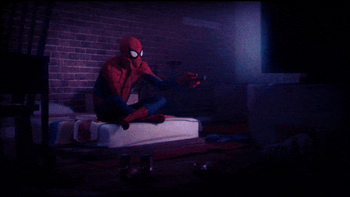 a man in a spiderman costume is sitting on a bed holding a remote control