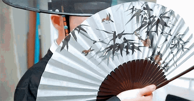 a man wearing a hat is holding a fan with a painting of birds on it .