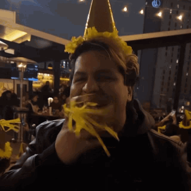 a man wearing a party hat with yellow flowers on it eating yellow noodles