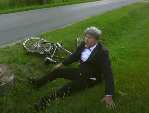 a man in a tuxedo is laying in the grass with a bicycle on the side of the road