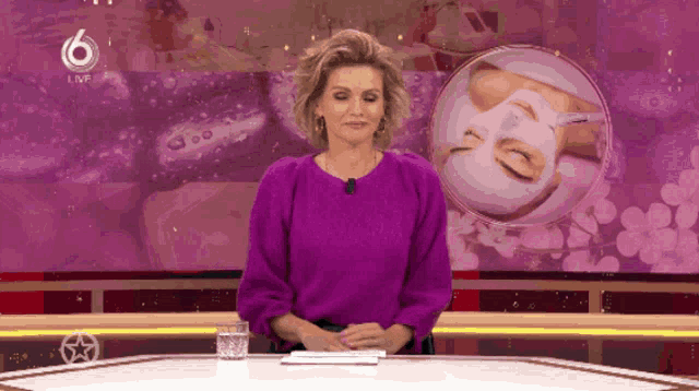 a woman in a purple sweater is sitting at a desk with a glass of water in front of a screen that says 6 live