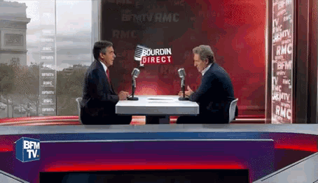 two men are sitting at a table with microphones in front of a sign that says bourdin direct on it