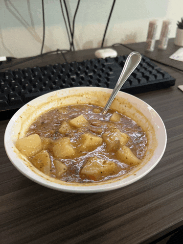 a bowl of soup with a spoon in it sits on a wooden table in front of a keyboard