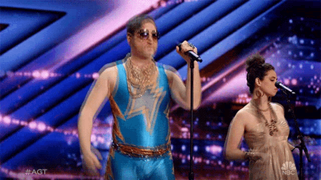 a man in a blue leotard is singing into a microphone while a woman sings behind him .