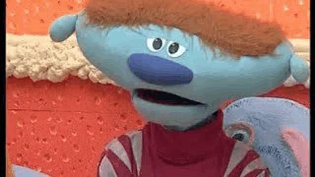 a blue puppet with a mustache and a red sweater is standing in front of a wall .