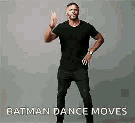 a man in a black shirt is dancing like batman and giving the middle finger .