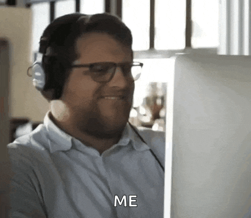 a man wearing headphones and glasses is sitting in front of a computer screen and saying `` me '' .
